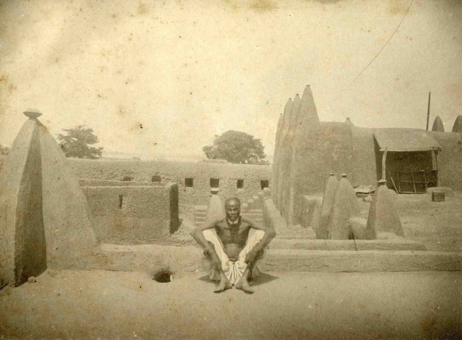un homme assis devant un habitat traditionnel en terre.