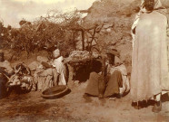 Devant un gourbi (habitat en torchis), une femme pilant, deux autres travaillant la laine, un berceau suspendu.